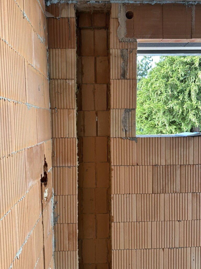 Rohbau aus Backstein, vertikale Wandöffnung, Blick nach draußen auf grüne Bäume.