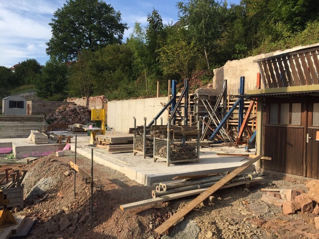 Baustelle mit Gerüst, Bauholz, Betonplatten und Kies im Vordergrund; rechts eine Holztür.