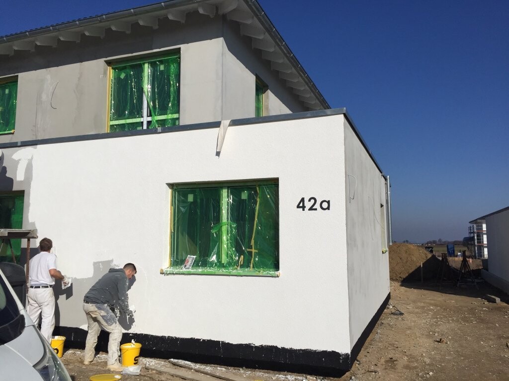 Weiße Neubauwand mit grün abgedeckten Fenstern; Bauarbeiter arbeiten am Rohbau.