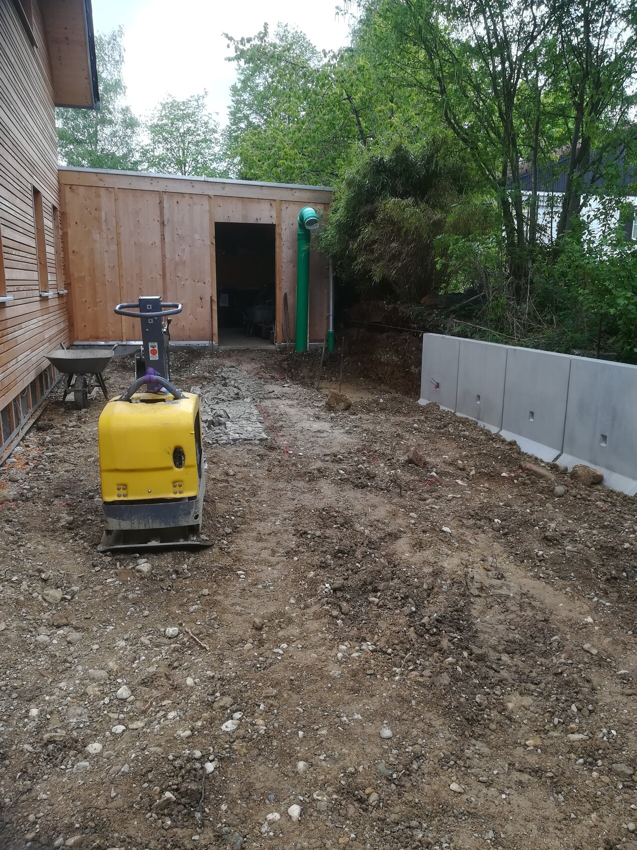Hinterhof-Baustelle: Gelber Plattenverdichter auf unbefestigtem Boden neben Holzschuppen
