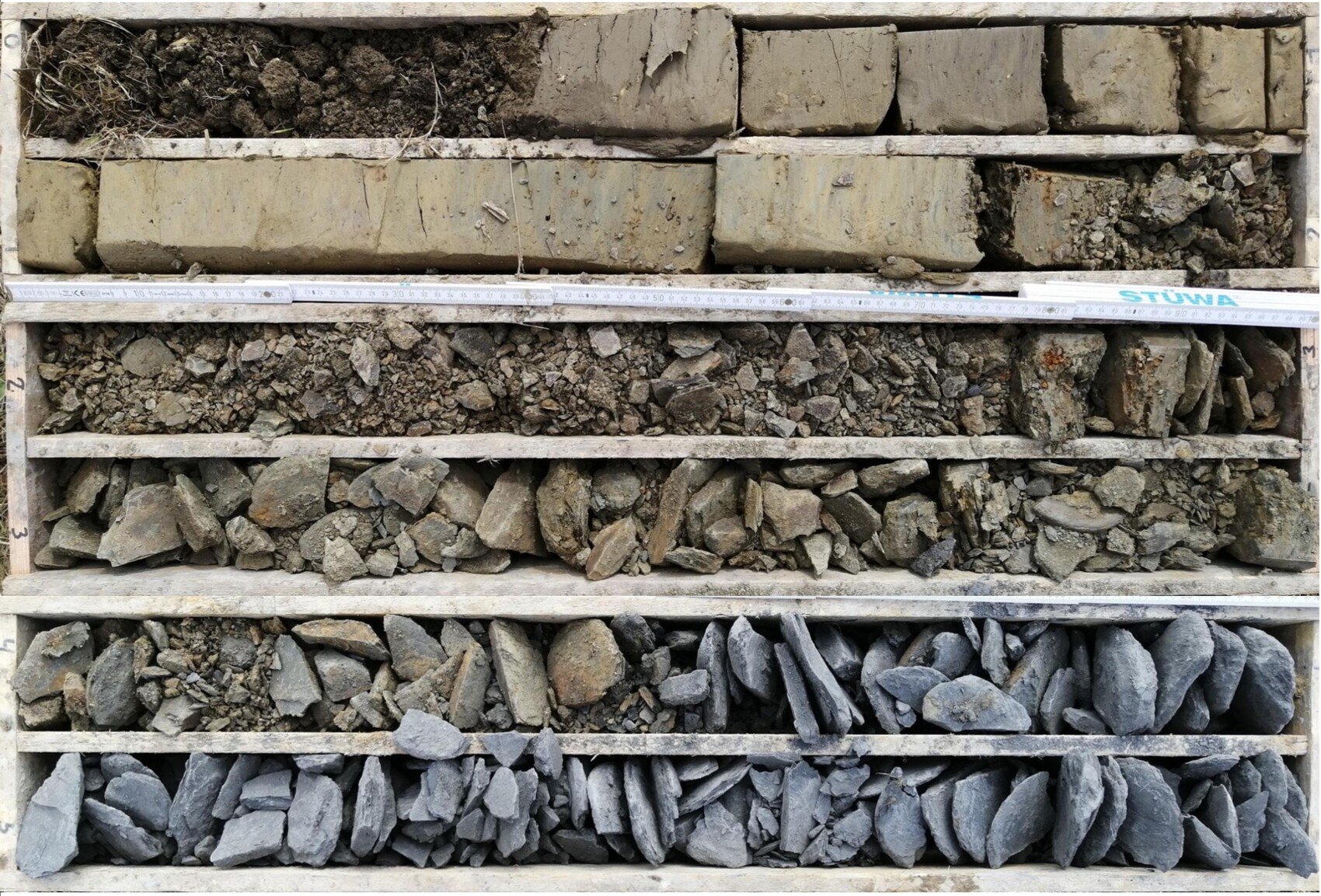 Holzkiste mit Gesteinsproben in Fächern: Erde, Kies, Bruchstücke und Steine; Messband darüber.