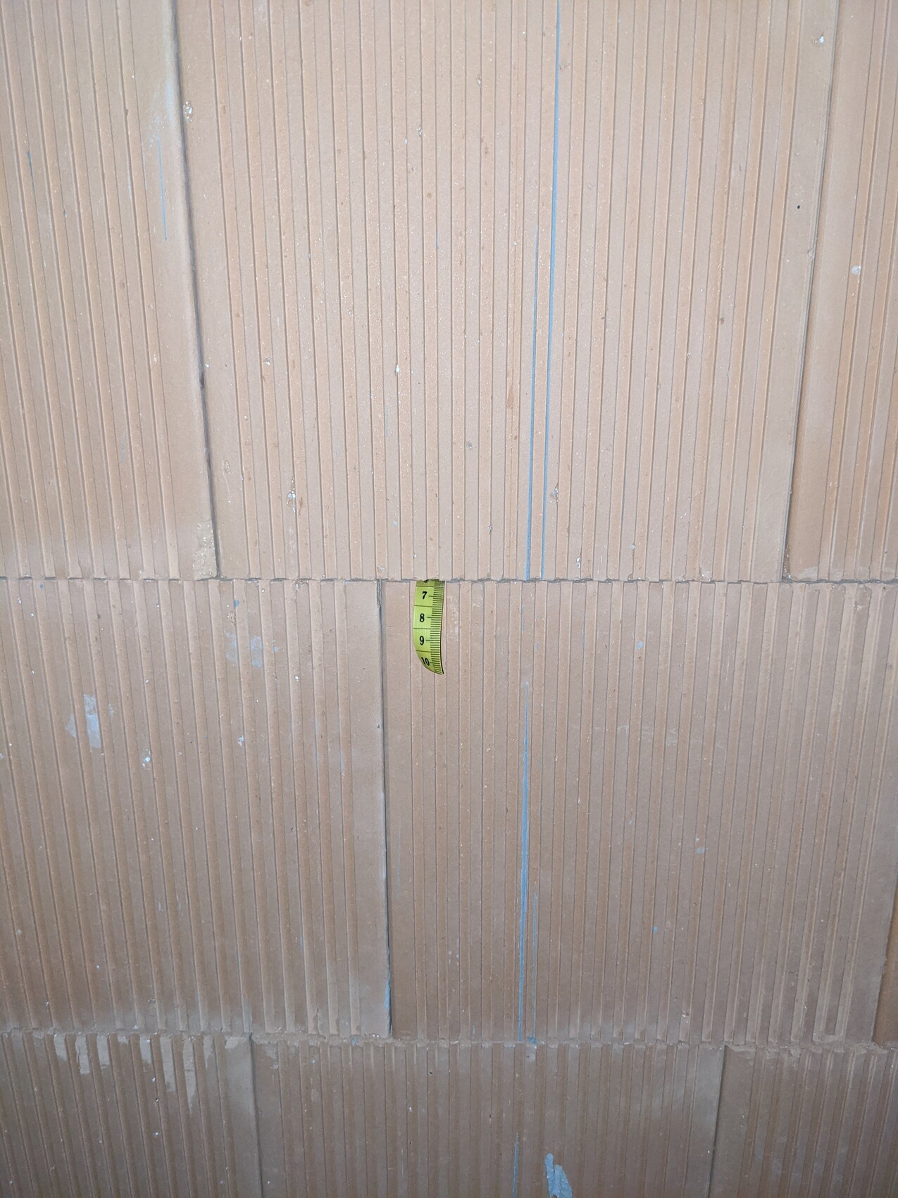 Braune Wand mit vertikalen Rillen; ein gelbes Maßband hängt mittig als Maßstab.