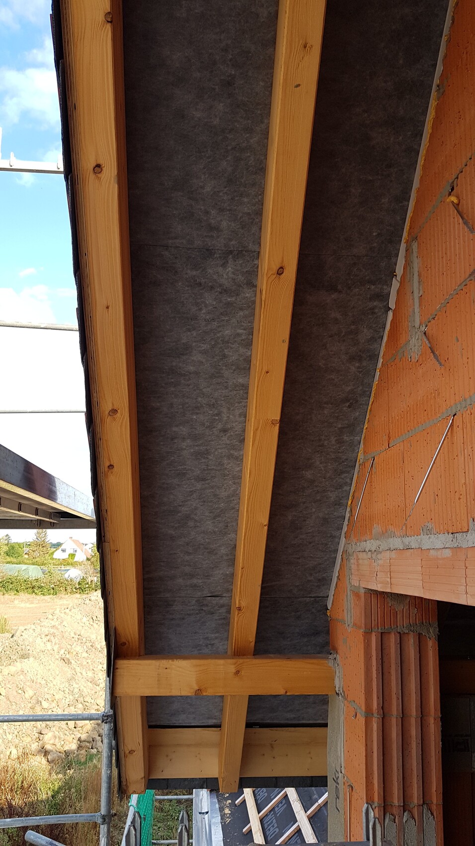 Dachkonstruktion mit Holzsparren, schwarzer Dämmung und Ziegelwand auf Baustelle.