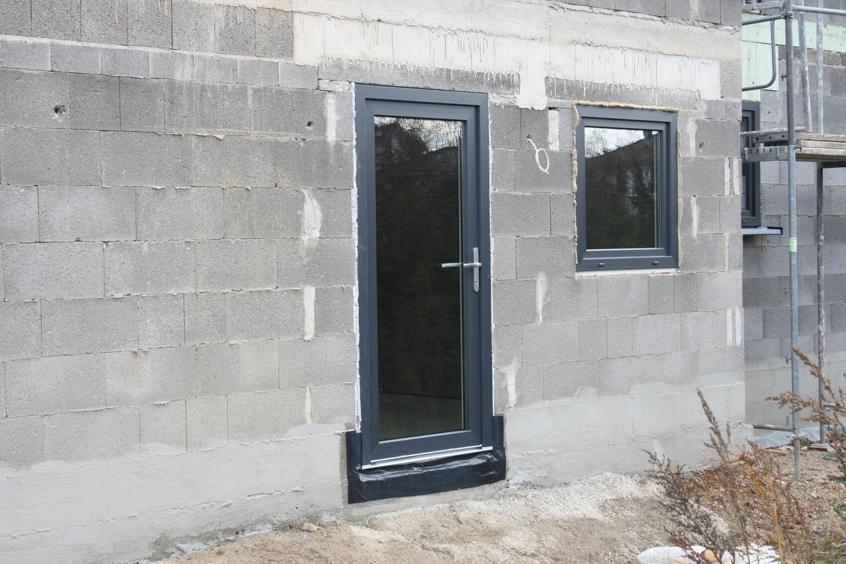Baustelle am Rohbau: Betonblöcke, Glastür und Fenster, rechts Baugerüst.