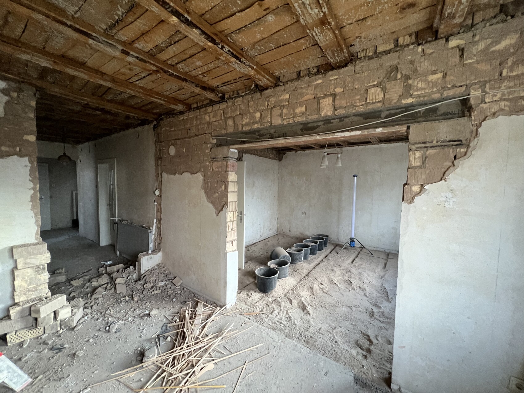 Rohbauzimmer mit freigelegten Ziegelwänden, Holzbalkendecke und Trümmern; Eimerreihe im Raum