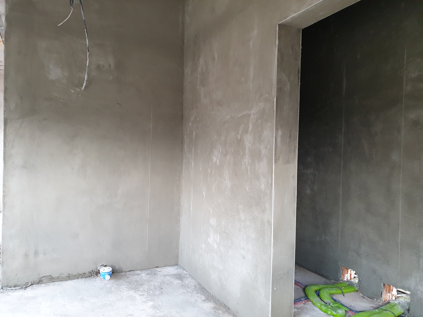 Rohbau-Raum aus Beton, offenes Durchgangsportal, lose Kabel, Eimer links unten, grüne Rohre rechts.