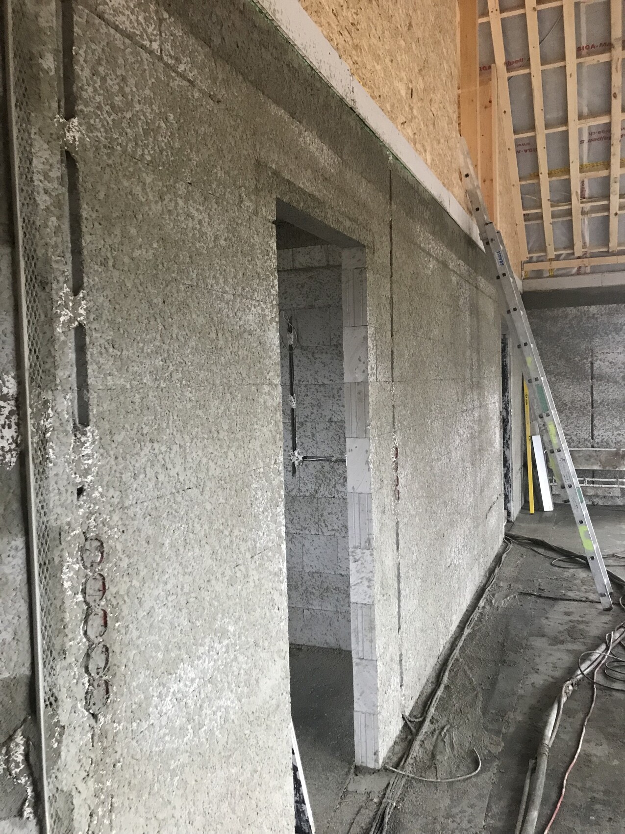 Innenraum einer Baustelle: raue Betonwand, Türöffnung, aufgestellte Leiter und Kabel am Boden.