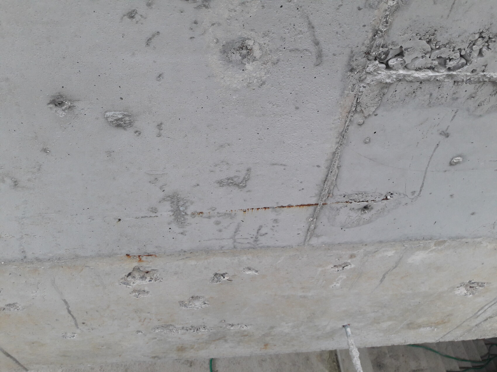 Betonoberfläche mit Rissen, Flecken und rostigem Streifen entlang einer Naht; grüner Schlauch unten.