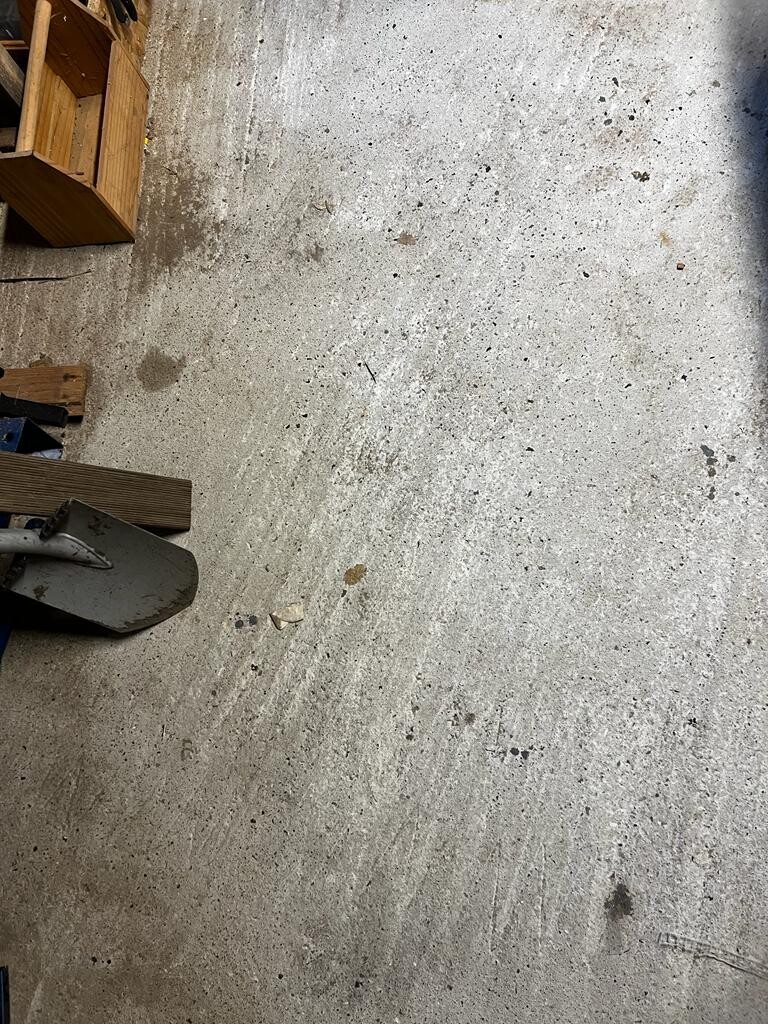 Betonboden mit Staub und Kieselsteinen; links Holzbretter, Schaufel am unteren Rand.