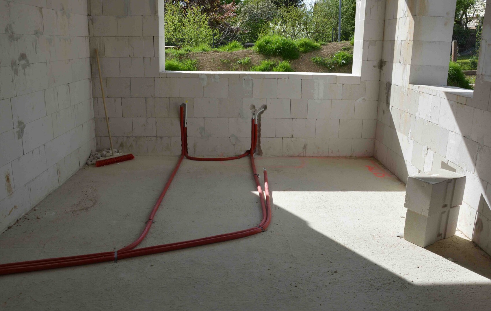Rohbau: Raum mit weißen Steinwänden, roten Kabelrohren am Boden undFenster mit Gartenblick.