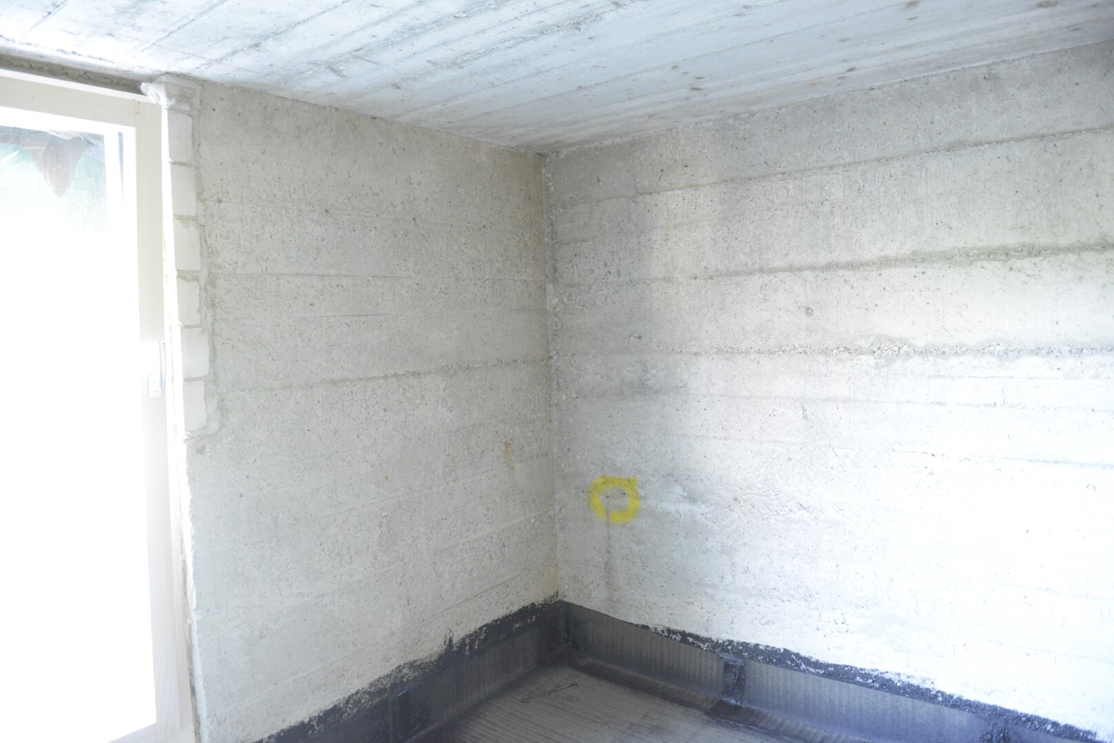 Keller-Eckraum aus Beton, gelbe Kreismarkierung an der Wand und dunkle Bodenabdichtung