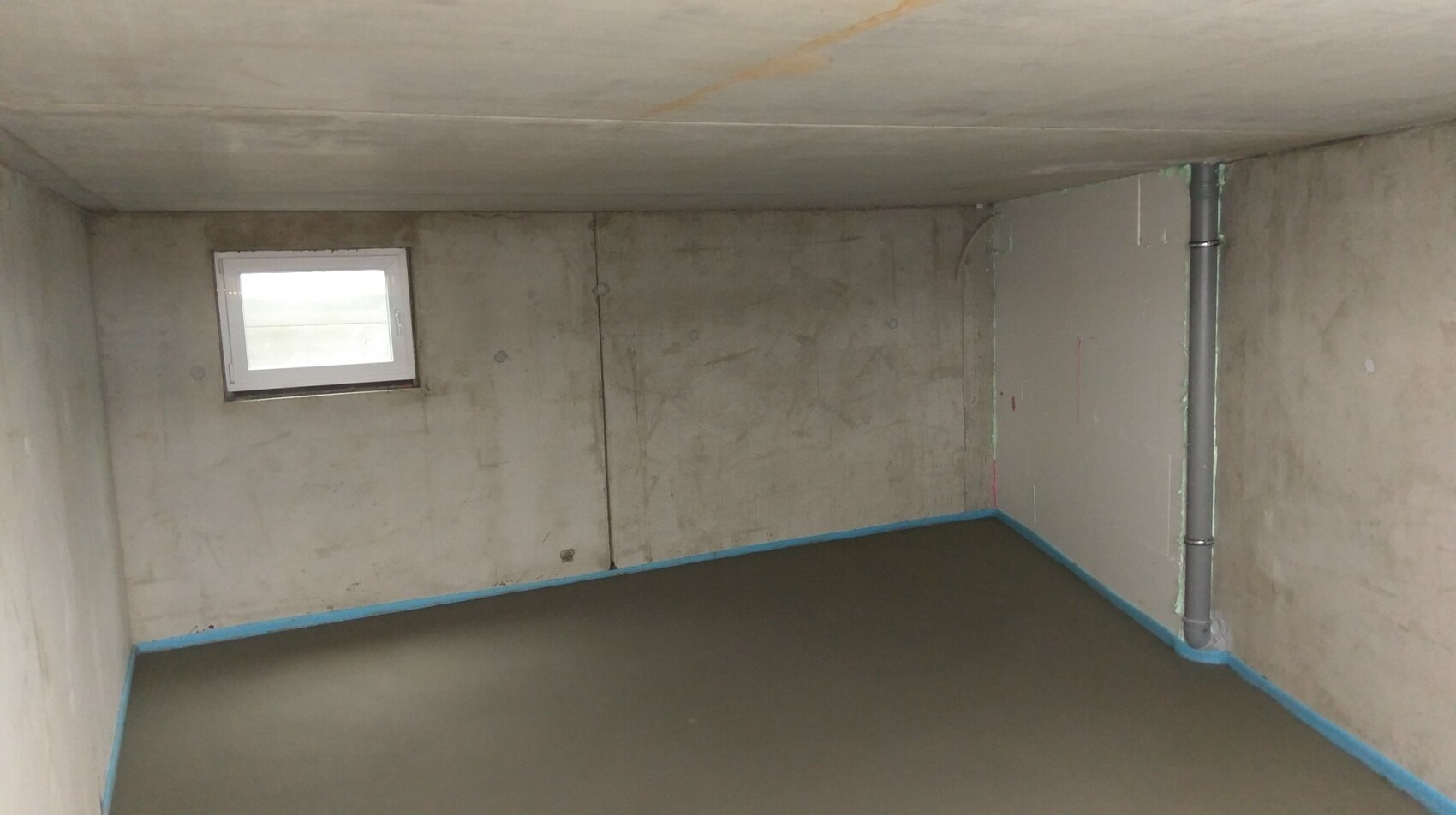 Kellerraum mit rohen Betonwänden, kleinem Fenster, Rohr und feuchtem Boden