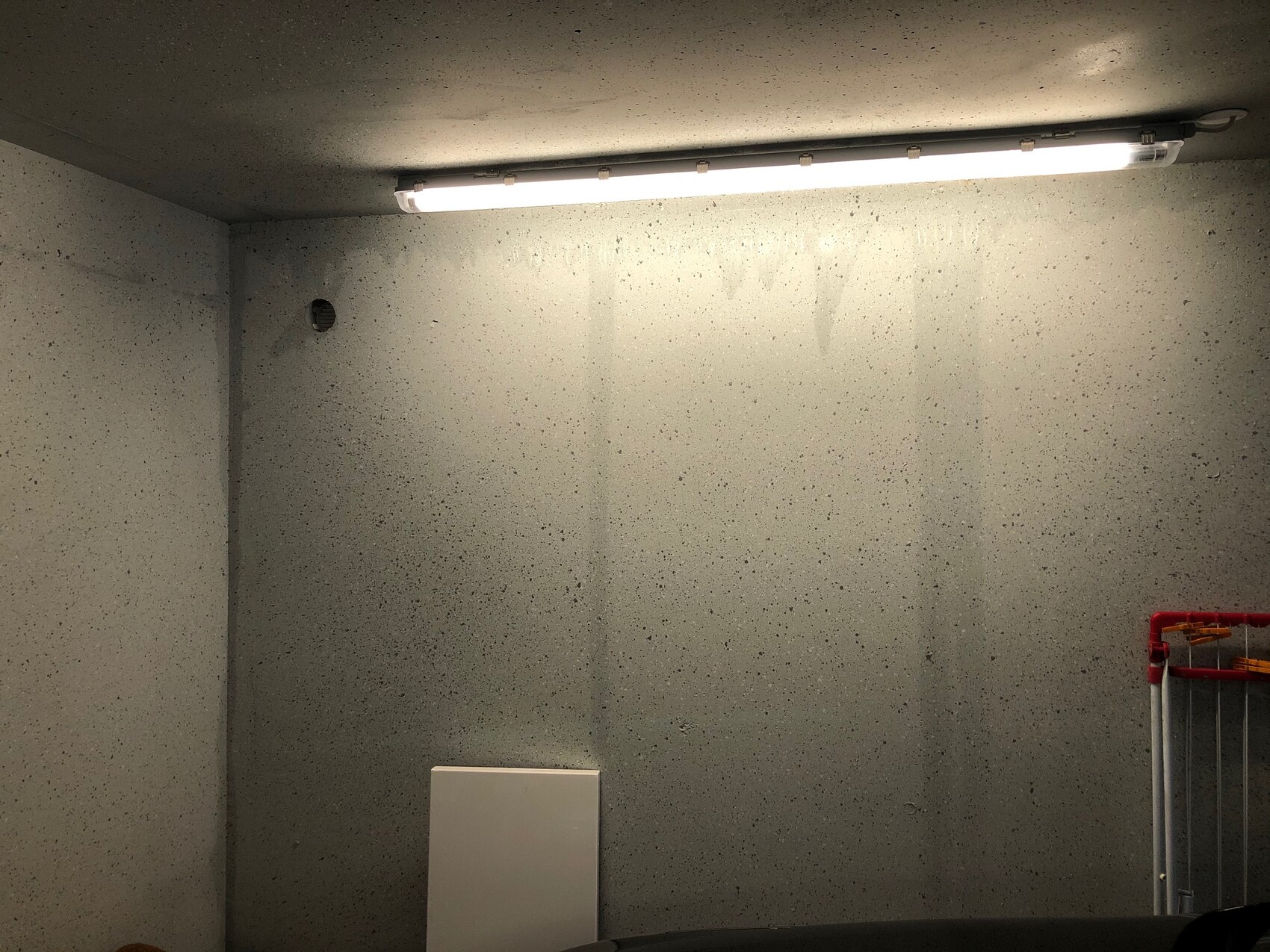 Betonraum/Garage mit Deckenleuchte, Wandloch links, weisses Brett, roter Kleiderstaender rechts.