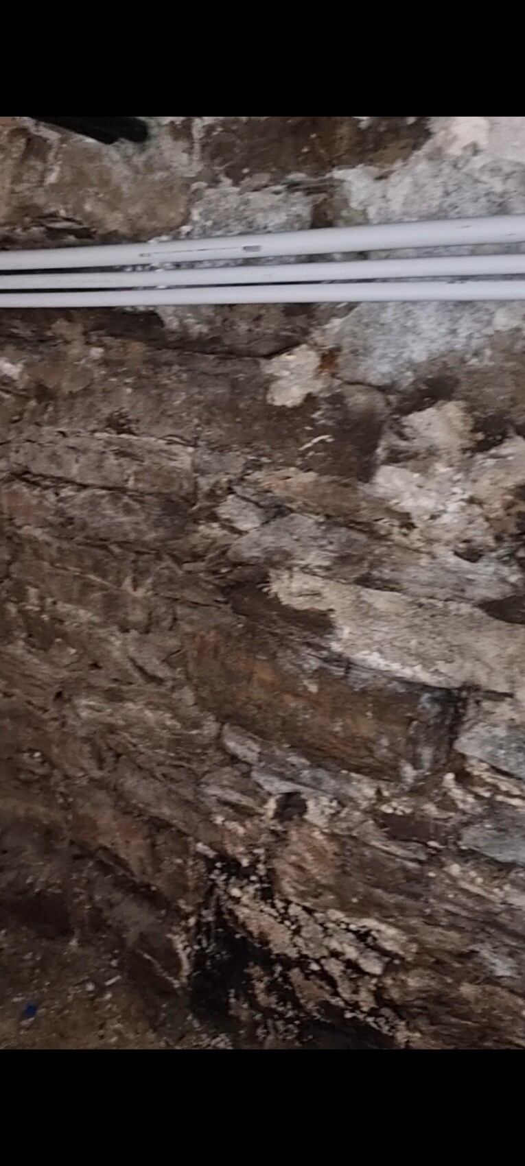 Unverputzte Steinwand mit mehreren weißen PVC-Rohren; feuchter, unregelmäßiger Boden.