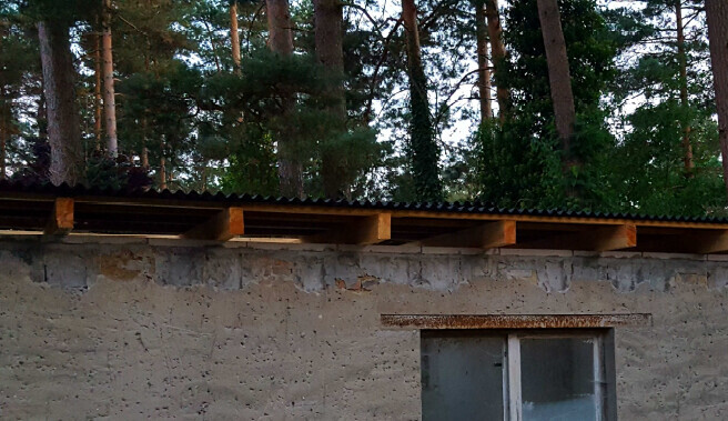 Außenwand eines unfertigen Hauses mit Fenster, Holzdach und Wald im Hintergrund.