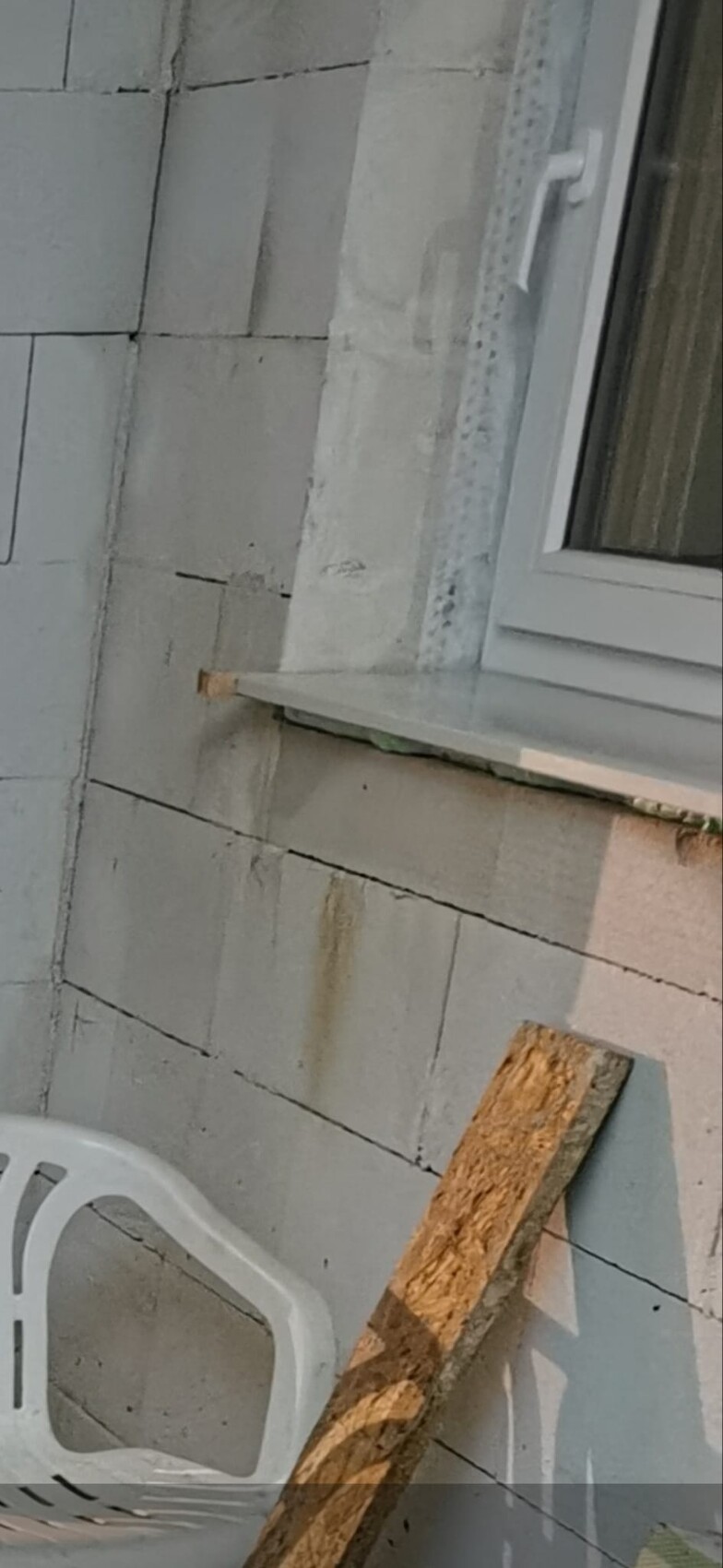 Außenwand aus grauen Betonblöcken mit Fenster; Holzbretter und Plastikstuhl sichtbar.