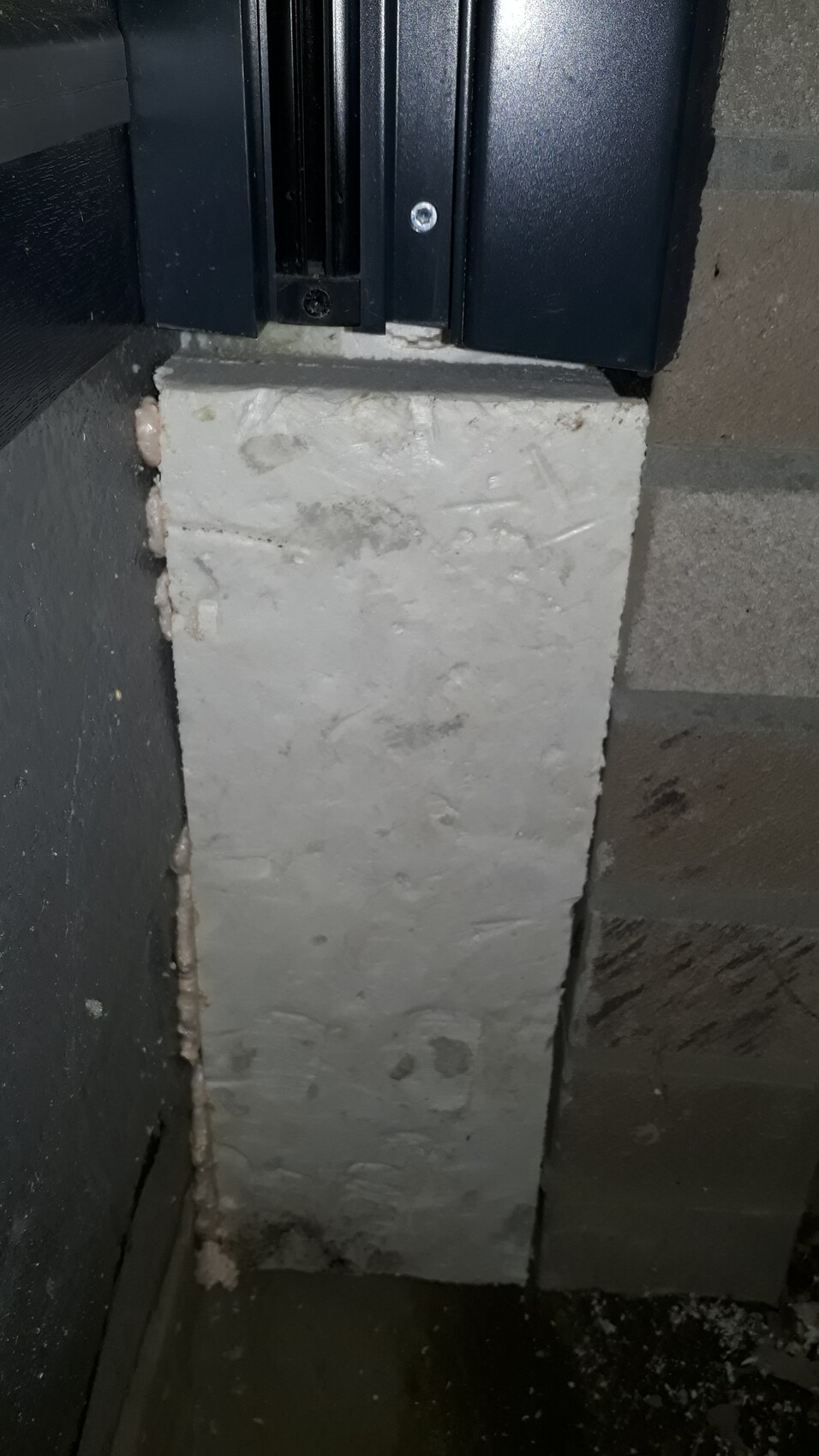 Grauer Betonblock in einer Ecke, links sichtbarer rosa Bauschaum; darüber schwarzes Metallteil.