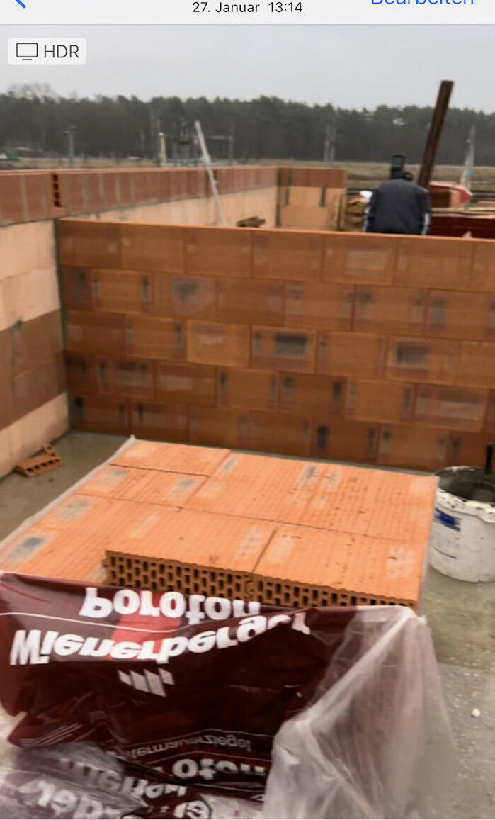 Baustelle mit orange Ziegelwänden, Ziegelstapel im Vordergrund, Bauarbeiter im Hintergrund.