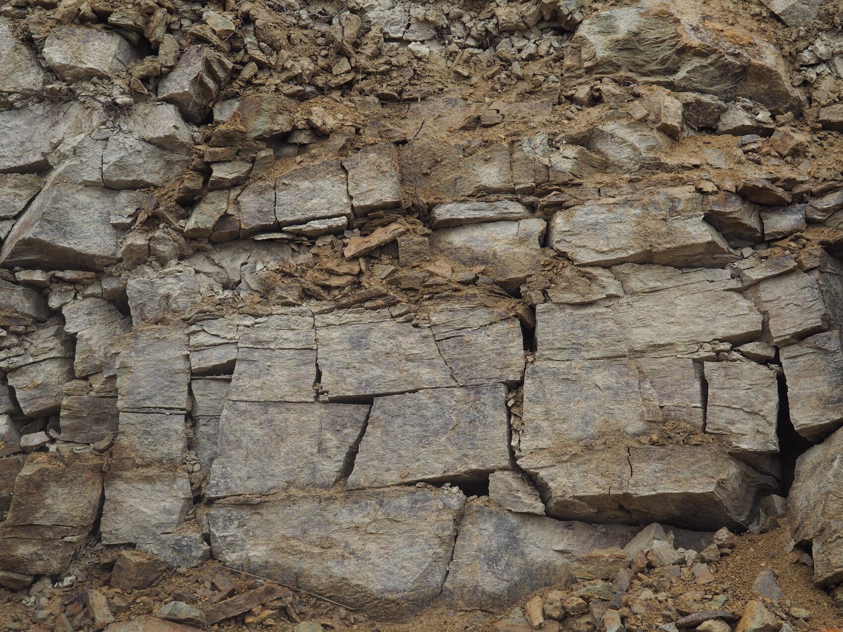 Steinmauer aus grob gestapelten, grau-braunen Blöcken mit Rissen und Erdkrümeln.