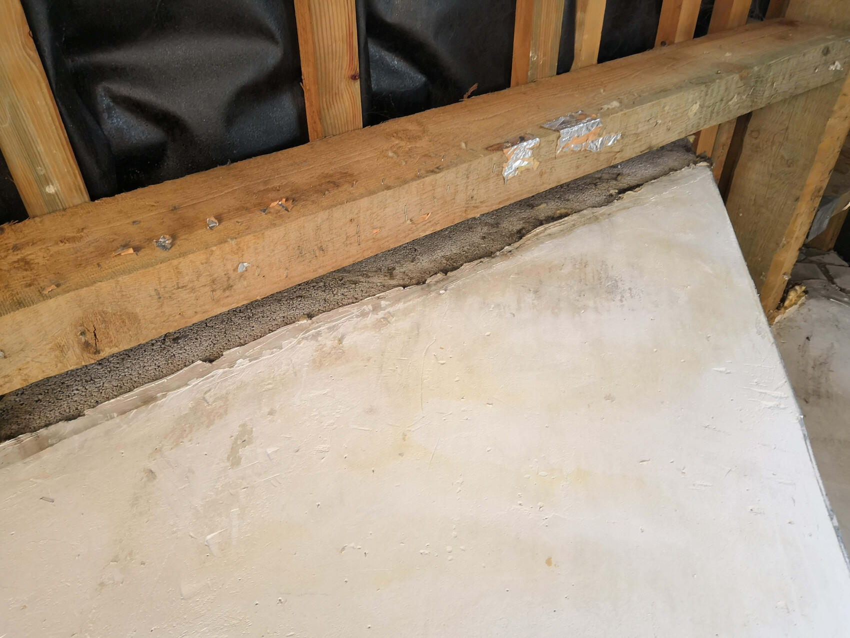 Baustelle: Holzständerwand mit schwarzer Dampfsperre; eine schräg stehende Betonplatte lehnt an der Wand.