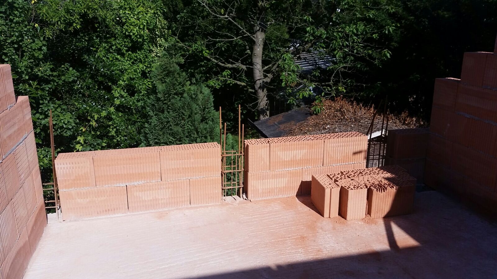 Baustelle mit roten Ziegelmauern, Bewehrungsstangen und Lochziegel-Stapeln vor grünem Hintergrund.