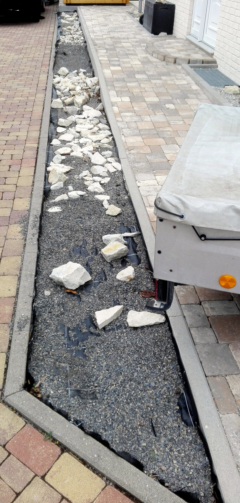 Langgestreckte Baugrube am Gehweg: Kies und Bruchsteine; daneben ein Bauanhänger.