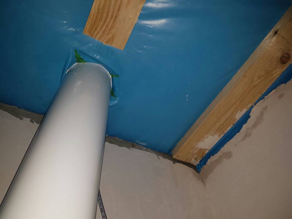 Großes weißes PVC-Rohr durch blaue Abdichtungsfolie im Keller, Holzbalken sichtbar