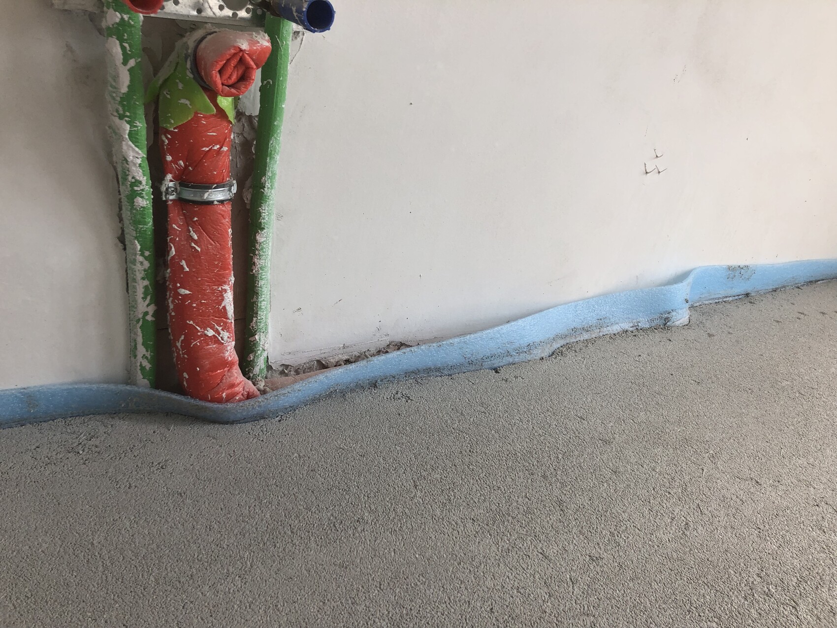 Rotes isoliertes Rohr neben grünen Rohren an einer Bauwand; blauer Bodenabschluss auf grobem Beton.