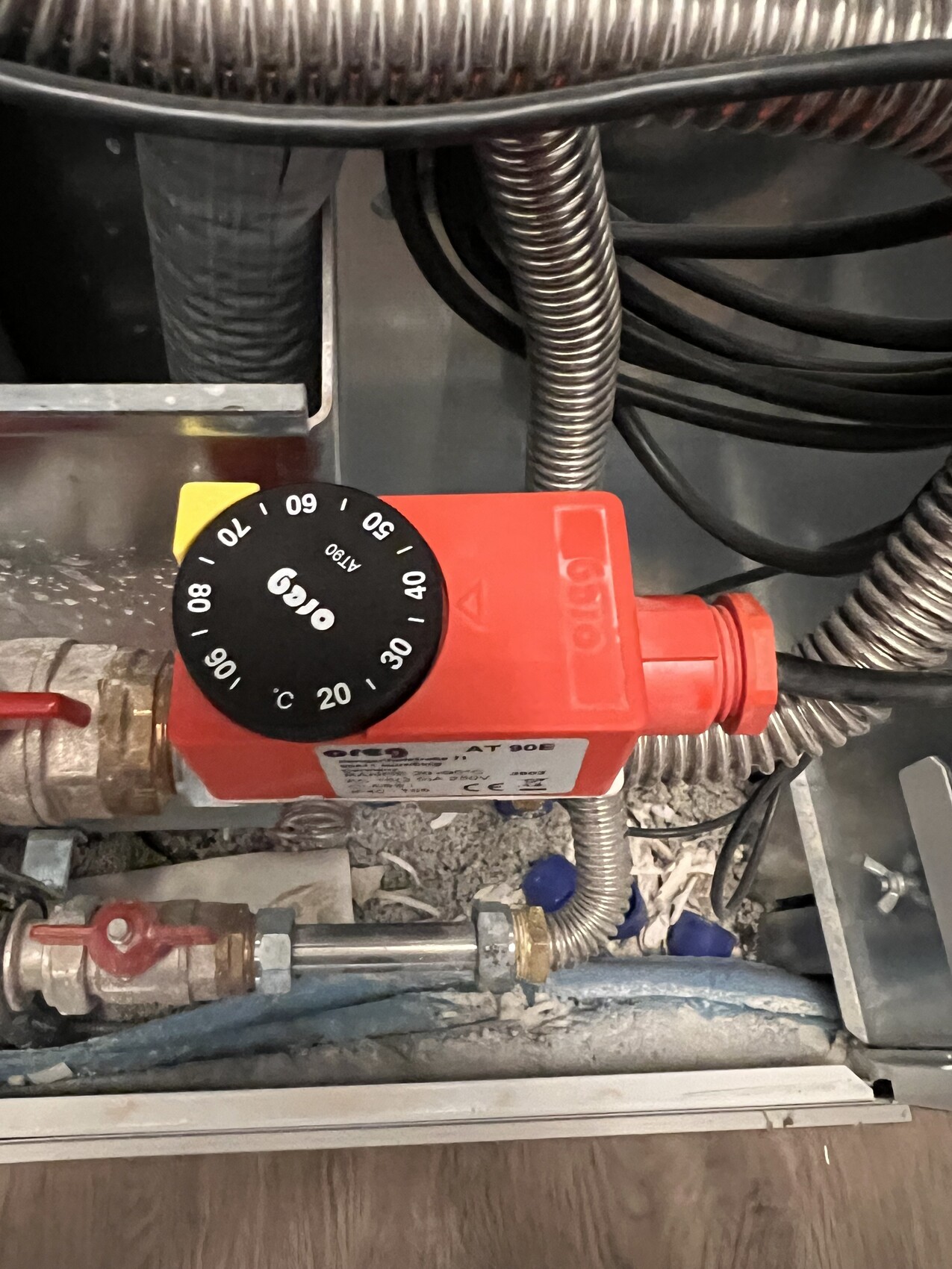 Roter Temperaturregler mit schwarzer Skala auf Heizungsrohr, umgeben von Kabeln und Metallleitungen.