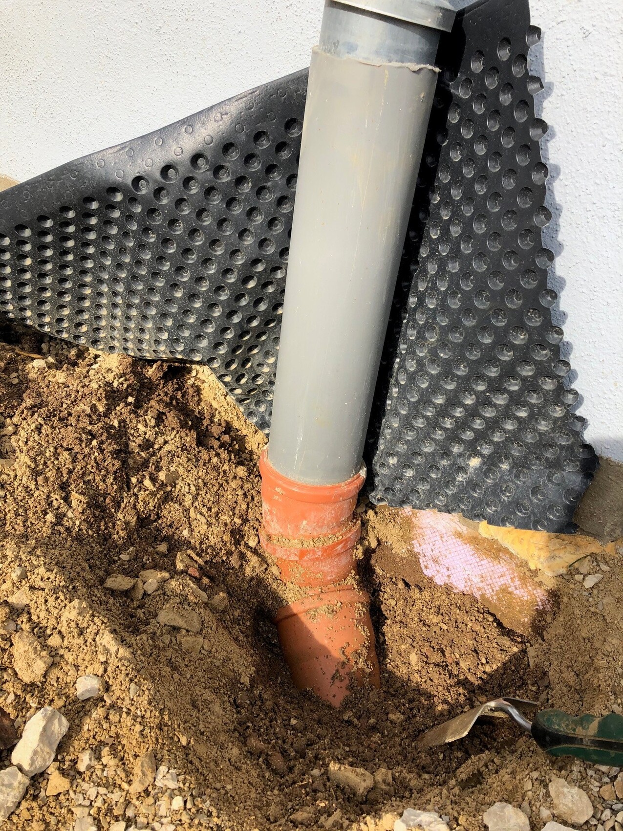 Baustelle zeigt graues Abflussrohr mit rotem Anschluss neben Fundamentwand, Drainagefolie.