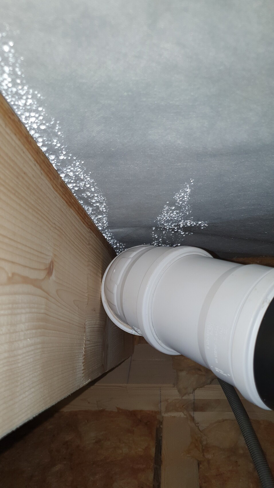 Weiße PVC-Rohrleitung mit Verbinder verläuft durch Holzrahmen neben silberner Dämmfolie.