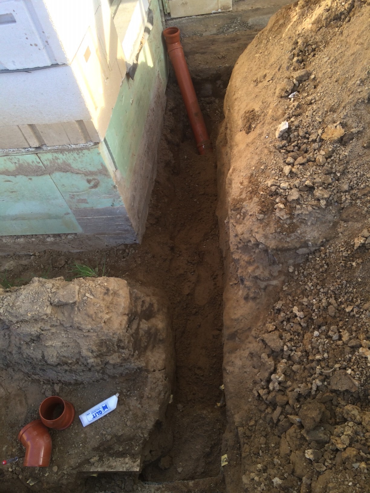 Graben neben dem Hausfundament mit rotem PVC-Abflussrohr im Erdreich.