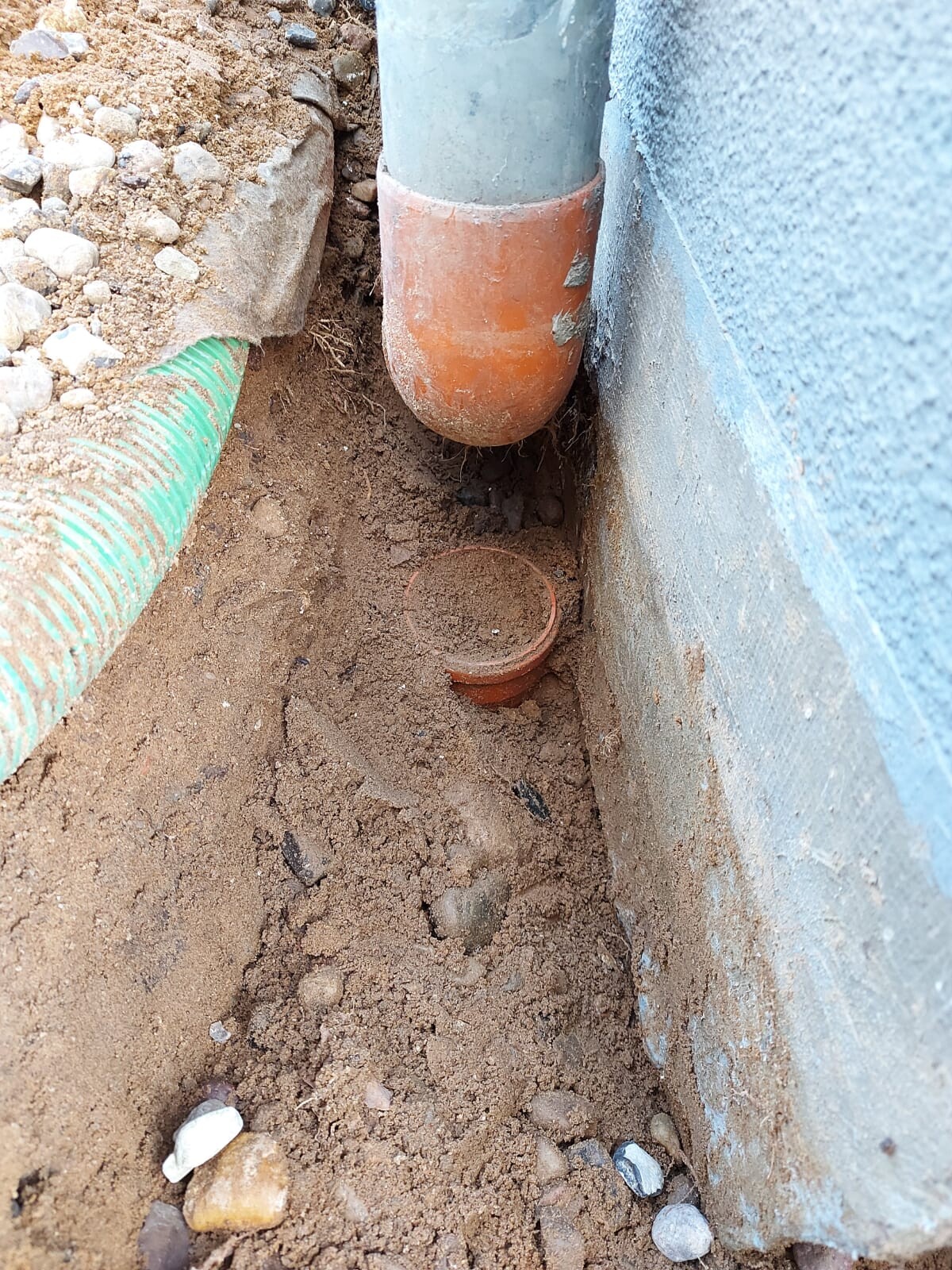 Kanalrohr unter der Wand; orangefarbener Anschluss, daneben grüner Schlauch im Sandboden.
