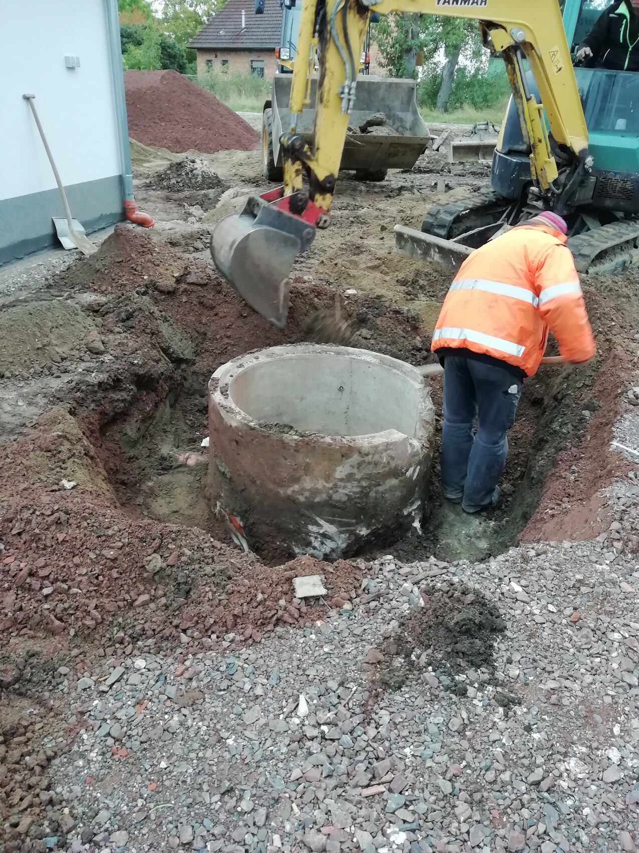 Bauarbeiter in orangefarbener Weste neben offener Baugrube; Bagger hebt Betonrohr zum Einbau.