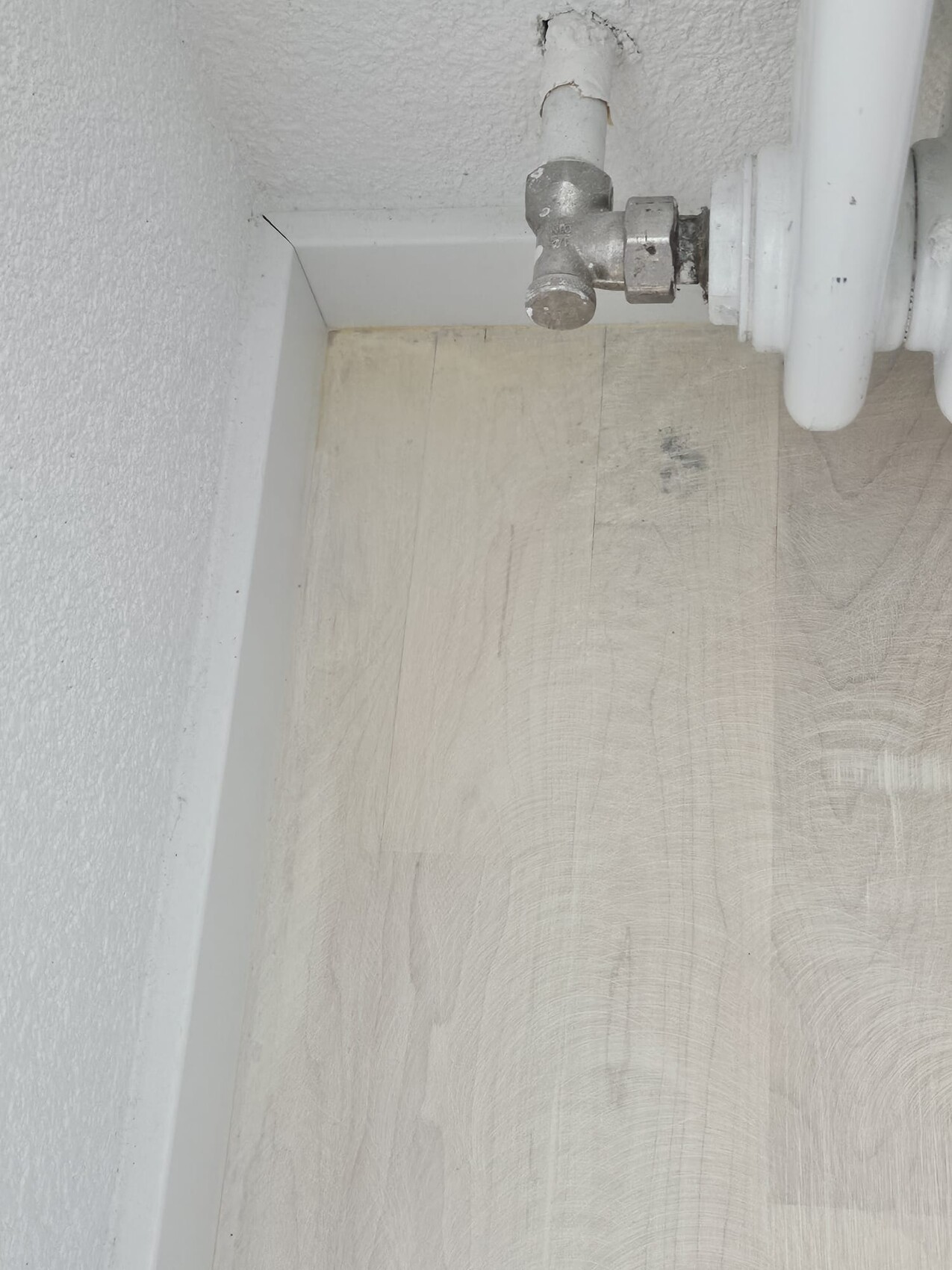 Eckansicht: Weißes Heizungsrohr mit Metallventil in der Ecke über dem Holzboden.