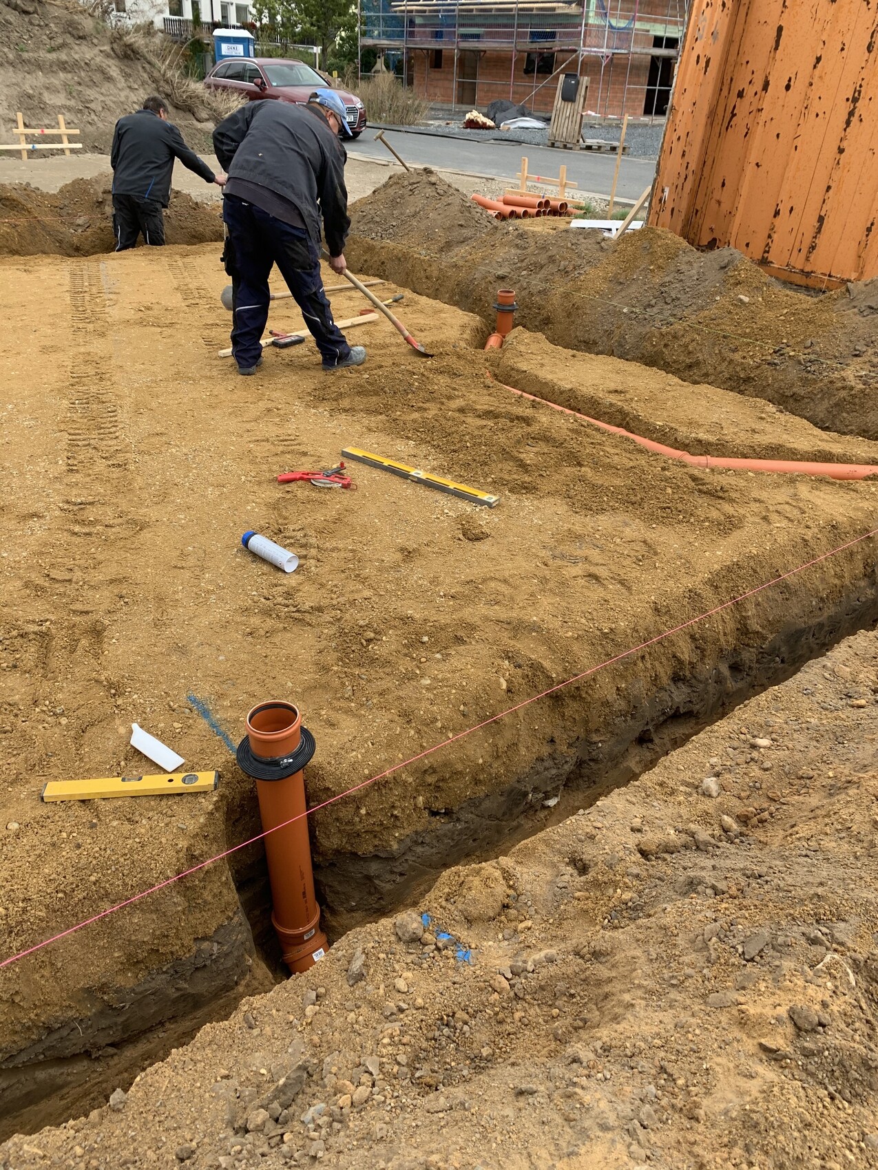 Bauarbeiter arbeiten in einer Baugrube und verlegen orange PVC-Rohrleitungen; Werkzeuge liegen bereit.