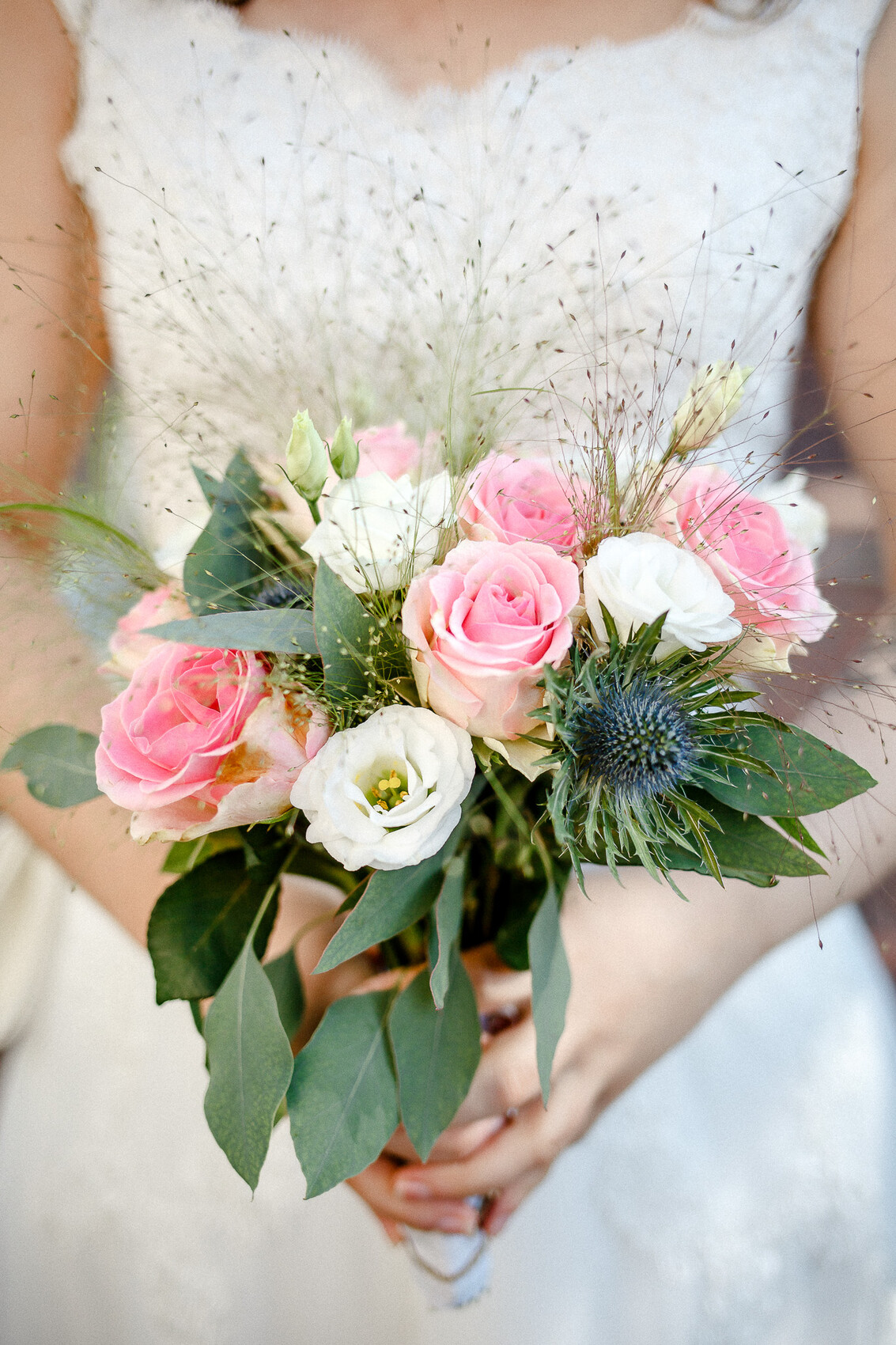 Braut in Hochzeitskleid hält Brautstrauß mit rosa Rosen, weißen Blüten, grünem Laub und Distel.