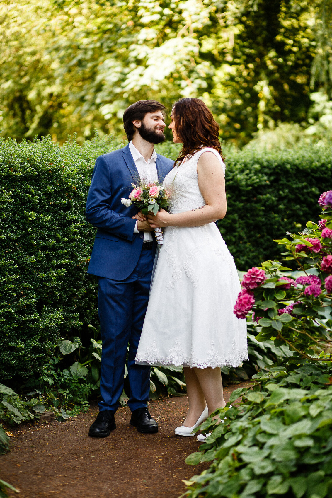 Hochzeitspaar im Garten: Braut in Weiß und Bräutigam in Blau schauen sich an, Braut hält Strauß.