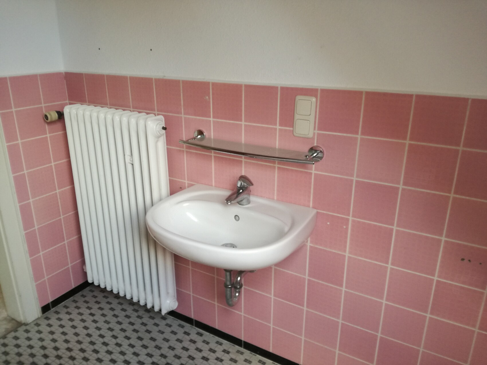 Waschbecken im Badezimmer mit Armatur vor rosa Fliesen, links Heizkörper und Handtuchhalter.