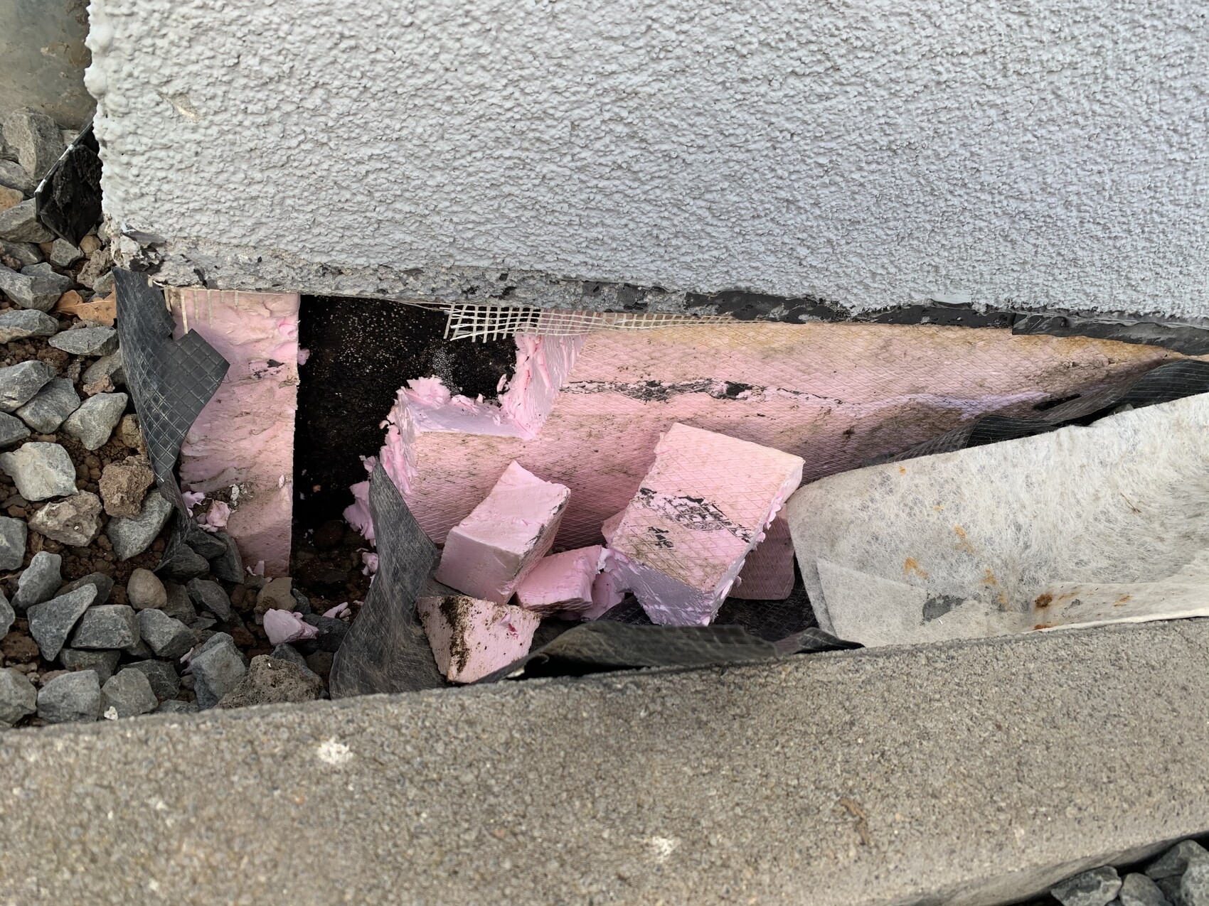 Fundamentunterkante eines Gebäudes: rosa Dämmplatten zerbrochen, schwarze Abdichtungsbahn, Kies.
