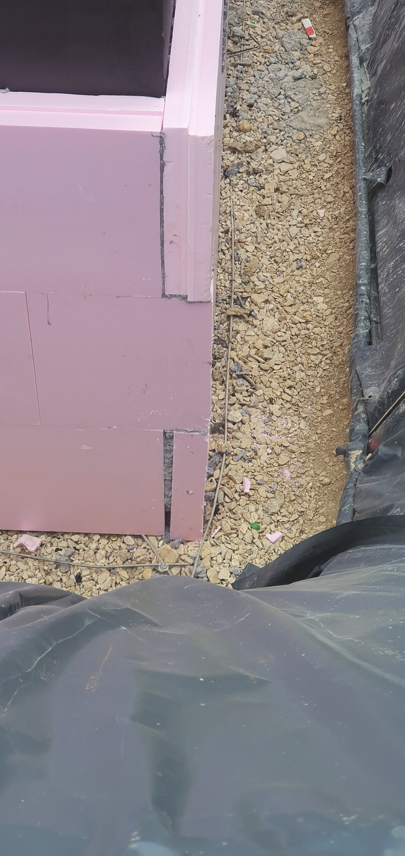 Baustelle mit rosa Dämmplatten an einer Wand, Kiesboden und schwarze Bauplane.