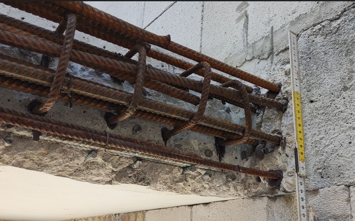 Rostige Bewehrungsstangen aus Stahl in einer Betonwand; rechts sichtbar ein Maßband.