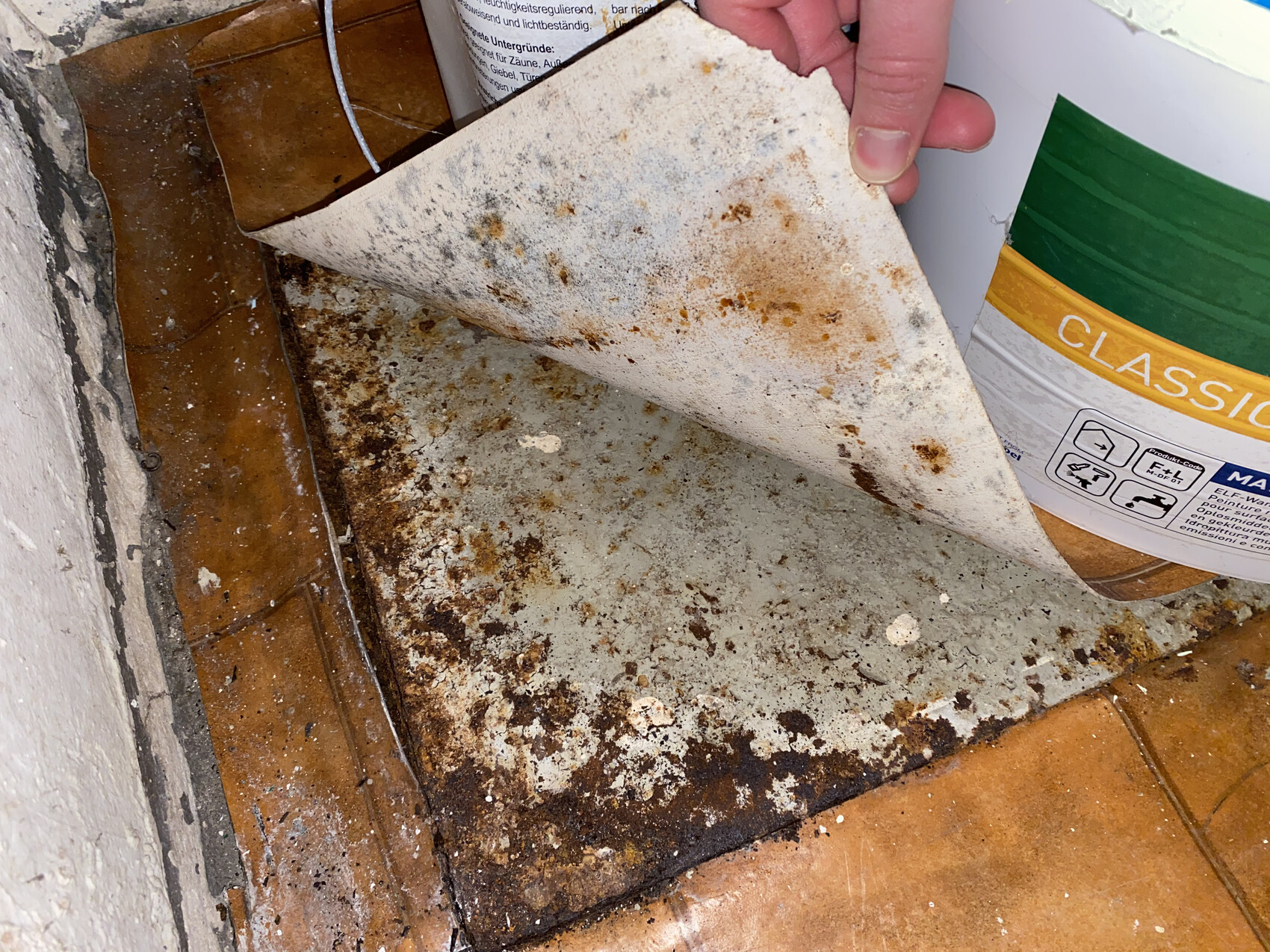 Hand hebt schmutzige, rostige Bodenfläche mit Flecken; daneben grün-gelber Farbeimer.