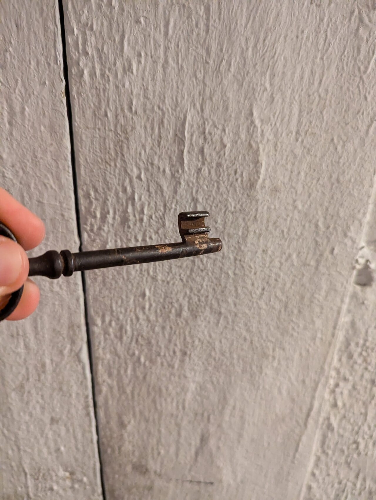 Hand hält rostigen, alten Schlüssel mit zwei Zinken vor rauer Holzwand.