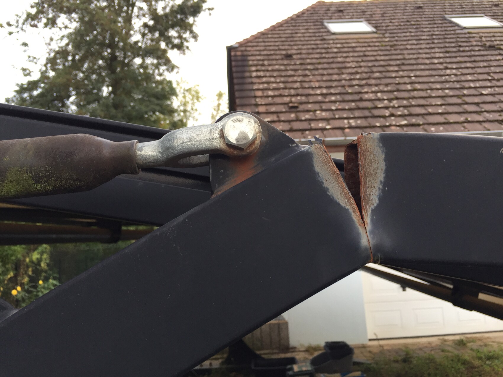 Rostige Stahlträger-Verbindung mit Riss, Bolzen sichtbar; Dach und Haus im Hintergrund