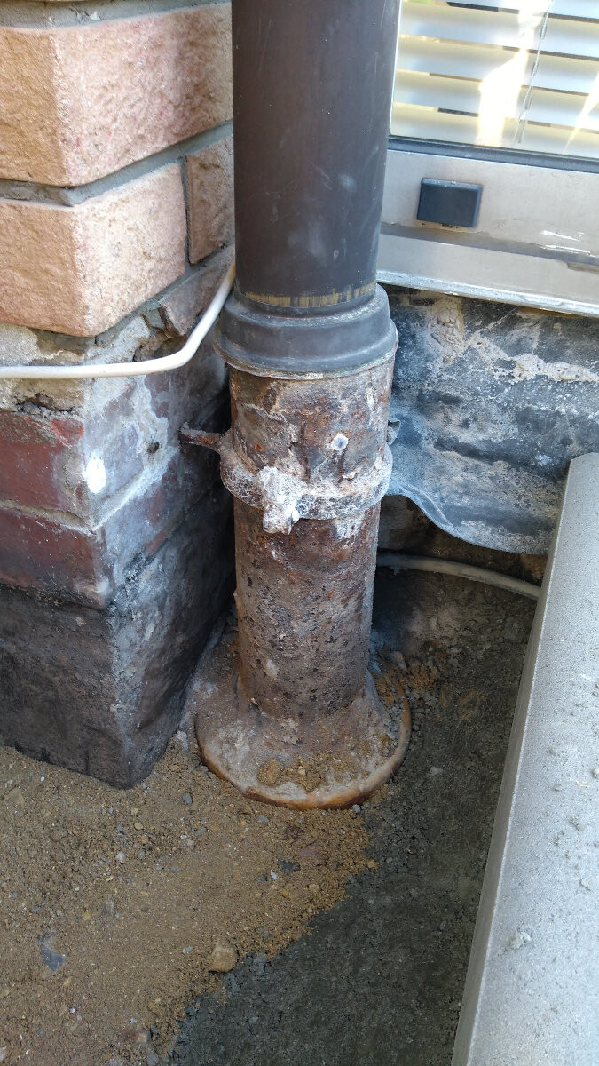 Rostiges Abwasser-/Abflussrohr am Fundament neben Backsteinwand; deutlich korrodiert.