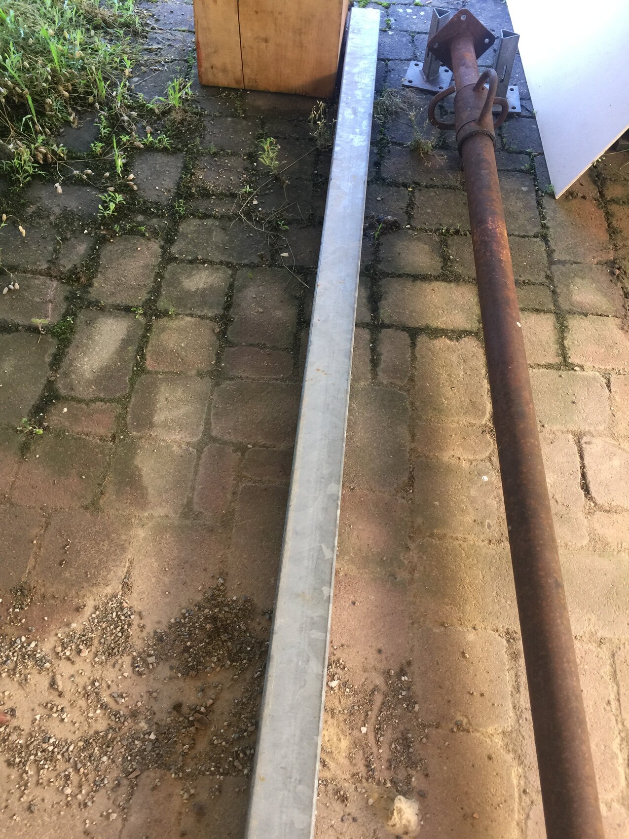 Rostiges Rohr mit Flansch liegt neben silbernem Metallprofil auf gepflastertem Boden.
