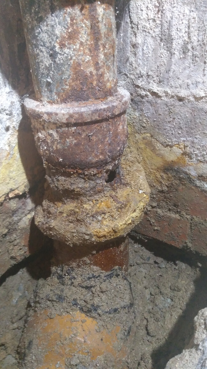 Rostiges Rohr mit Muffe, stark korrodiert, von Schlamm am Fundament umgeben.