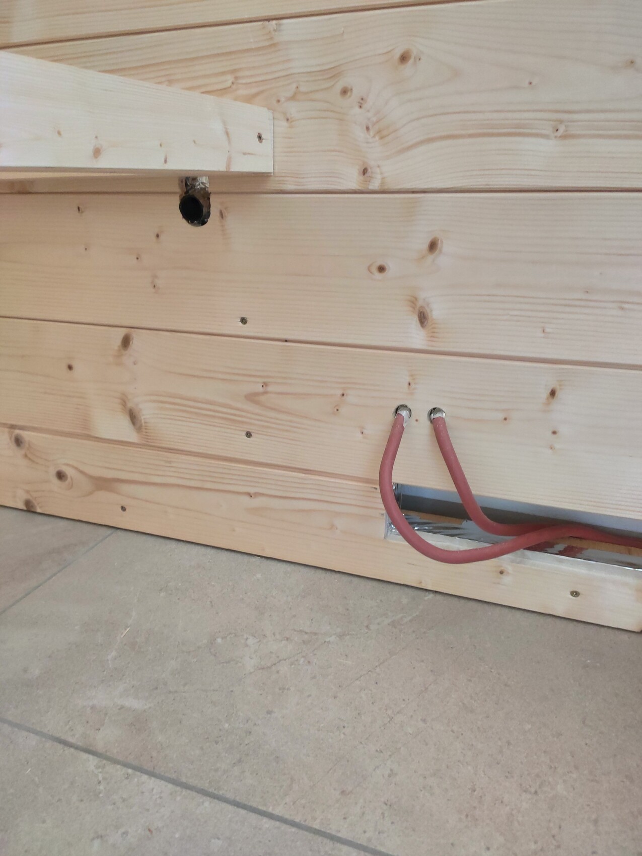 Zwei rote Kabel ragen aus einer Öffnung im unteren Holzpaneel der Wand; Betonboden darunter.