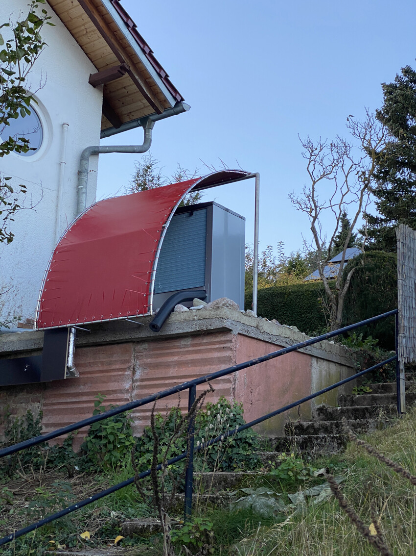 Rote halbrunde Stoffüberdachung über grauer Metallbox neben dem Haus; Gartenstufen sichtbar